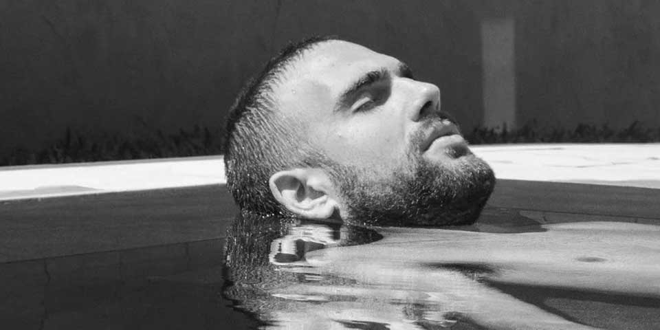 Mann mit Bart liegt mit geschlossenen Augen im Wasser, nur Kopf und Schultern schauen heraus
